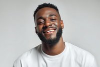 Happy young African American man with beard smiling with eyes closed wearing white t-shirt on gray background expressing joy and positive emotions.