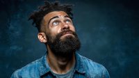 Young bearded man with curly hair looking up thoughtfully against dark blue background. Contemplative expression for inspiration and creativity concepts.