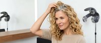 Attractive mature blonde woman with curly hair touching her head in modern bathroom mirror reflection showing confidence and self-care routine.