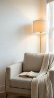 Cozy reading corner with beige armchair, knitted throw blanket, floor lamp and book in modern minimalist living room interior for home decor.