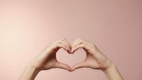 Hands forming a heart shape gesture on a soft pink background, symbolizing love, care, and positivity for Valentine's Day or wellness concepts.