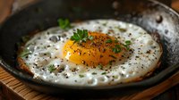 Sunny side up egg with black pepper and fresh parsley in cast iron skillet on wooden surface, bright orange yolk and crispy edges, close up view.