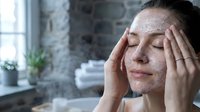Young woman applying facial scrub exfoliant with closed eyes in modern bathroom for skincare routine and beauty treatment at home.