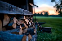 Stacked firewood in rustic outdoor shelter at sunset, with chopped logs neatly arranged against rural landscape background and green grass in foreground.
