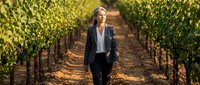 Professional blonde businesswoman walking through vineyard rows during golden hour sunset, corporate agriculture concept for wine industry marketing.