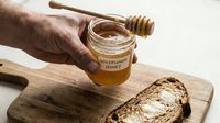 Male hand holding jar of wildflower honey with wooden dipper over cutting board with bread. Use for food, cooking, breakfast, natural products, organic ingredients, culinary content.