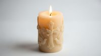 Cream colored pillar candle with ornate cross design burning with flame on white background for religious ceremonies and spiritual meditation.