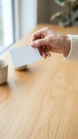 Senior woman holding blank white business card at wooden desk with coffee cup and plant, suitable for corporate branding and marketing materials.