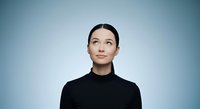 Young woman with East Asian ethnicity wearing black turtleneck against blue gradient background, looking upward with contemplative expression, studio portrait for corporate and lifestyle use.