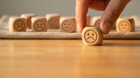 Hand selecting wooden cube with sad face emoji from row of happy face blocks on wood surface representing negative feedback choice and emotion selection.