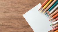 Colorful wooden pencils arranged on blank white paper against wooden desk background, creating diagonal composition with copy space for creative projects.
