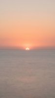 Serene ocean sunset with golden sun descending toward calm water horizon, soft peachy and pink sky gradients creating tranquil seascape for relaxation and travel content.
