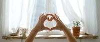 Hands forming heart shape in bright bedroom with white curtains and natural sunlight creating romantic morning atmosphere for wellness and self-care concepts.