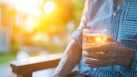Glowing golden beer in glass held against sunset light, creating atmospheric bokeh effect with warm sunbeams filtering through refreshing beverage.