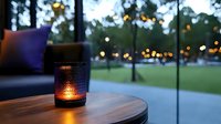 Cozy evening atmosphere with glowing candle lantern on wooden table at outdoor patio, string lights and green park in blurred background. Perfect for lifestyle content.