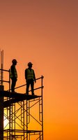 Two construction workers in safety vests and hard hats standing on scaffolding silhouetted against dramatic orange sunset sky for industrial projects.