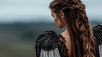 Young Caucasian woman with long braided hair wearing black lace and feather wings against moody nature background, fantasy fashion portrait in dark atmospheric style.