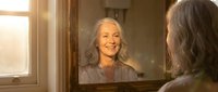 Mature woman with gray hair smiling at her reflection in bathroom mirror during morning routine, self-care and positive aging concept.