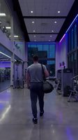 Athletic Caucasian man walking through modern gym with purple LED lighting carrying sports bag during evening workout session in fitness center.