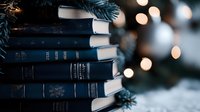 Stack of vintage navy blue books with gold lettering against blurred Christmas lights and pine branches creating cozy winter holiday atmosphere with bokeh effect.