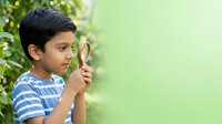 Young boy with dark hair and South Asian ethnicity, wearing blue striped shirt, holding magnifying glass outdoors among green foliage. Suitable for education, exploration, discovery, and learning concepts.
