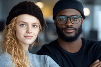Young diverse couple portrait with blonde woman in knit beanie and African American man in glasses and cap smiling together in modern indoor setting.