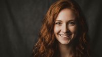 Young redhead woman with curly hair smiling warmly at camera against dark textured background. Natural portrait photography for lifestyle content.