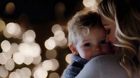Young blonde mother tenderly embracing small child against blurred bokeh lights background, intimate family moment capturing maternal love and holiday atmosphere.