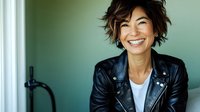 Young Asian woman with short tousled hair wearing black leather jacket and white t-shirt laughing genuinely while sitting on couch, expressing joy and confidence.