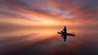 Silhouette of person in kayak on calm water during golden sunset with dramatic orange and pink sky clouds reflecting on mirror-like lake surface for peaceful recreation.