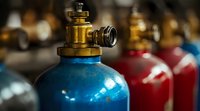 Close up view of blue industrial gas cylinder with brass valve fitting and regulator against blurred background of red tanks, selective focus on metallic details.