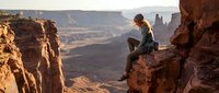 Young woman hiker sits on red rock cliff edge overlooking desert canyon landscape during golden hour sunset, perfect for outdoor adventure and travel content.