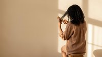 Young African American woman in cozy beige sweater touching forehead in contemplative pose with warm natural lighting and soft shadows on neutral wall.