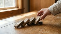 Caucasian male hand stopping falling wooden dominoes on timber surface near window, representing risk management and business prevention strategy.
