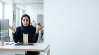 Middle Eastern woman wearing black hijab and blazer sitting at desk with hands raised in frustrated gesture, office environment with colleagues in background.