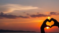 Hands forming heart shape silhouette against golden sunset sky over water. Romantic love symbol gesture at dusk with warm orange and pink clouds.
