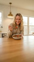 Happy blonde woman enjoying healthy breakfast bowl with berries in modern kitchen. Smiling female eating nutritious meal at home counter during morning routine.