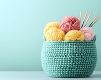 Cozy knitted mint basket filled with pink and yellow yarn balls and wooden knitting needles on pastel background, crafting supplies for DIY projects.