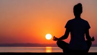 Young woman practicing yoga meditation in lotus pose silhouette against golden sunset over calm ocean water for mindfulness wellness and spiritual balance.