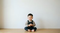 Young Asian boy, approximately 2-3 years old, sitting cross-legged on wooden floor against white wall, wearing casual brown and black clothing, holding gaming controller with happy expression.