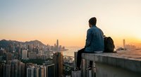 Young man sitting on rooftop edge overlooking modern city skyline at golden hour sunset with backpack, urban exploration and travel adventure concept.