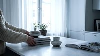 Woman in cozy sweater arranging books and coffee cups on white table by window in modern minimalist living room interior for home decor and lifestyle.