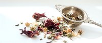 Loose dried herbal tea blend with rose petals, chamomile flowers and botanicals scattered on white surface next to silver mesh infuser strainer for brewing.