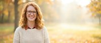 Happy young Caucasian woman with curly red hair and glasses smiling in autumn park with golden sunlight and warm bokeh background for lifestyle content.