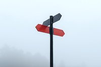 Minimalist directional sign post with red and gray arrows against foggy sky background, representing choice, direction and decision making concepts.