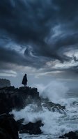Silhouetted figure standing on rocky coastline during dramatic storm with turbulent waves and dark swirling clouds overhead, conveying solitude and contemplation.