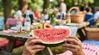 Close-up of hands holding a fresh watermelon slice at an outdoor summer picnic gathering with blurred people and food in background.