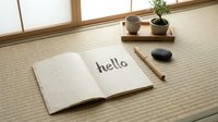 Open notebook with hello written on textured paper, placed on tatami mat in a minimalist Japanese room with bonsai and stone decor.