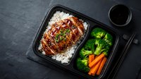 Teriyaki chicken rice bowl with steamed broccoli and carrots in black takeout container, healthy Asian meal prep lunch box on dark slate background.