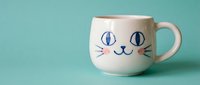 White ceramic mug with cute cat face design featuring blue eyes and whiskers on turquoise background for kitchen decor and beverage enjoyment.